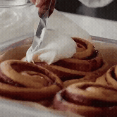 A Person Is Spreading Frosting On Cinnamon Rolls In A Pan . GIF