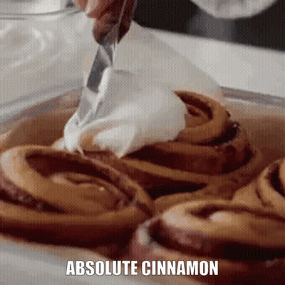 A Person Is Spreading Whipped Cream On Cinnamon Rolls With The Words Absolute Cinnamon Below Them . GIF