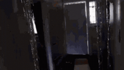 A Person Is Standing In A Dark Hallway Looking Out A Doorway . GIF