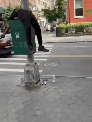 A Person Is Standing On A Green Trash Can On A Street GIF