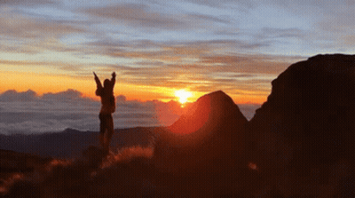 A Person Is Standing On Top Of A Hill With Their Arms In The Air At Sunset GIF