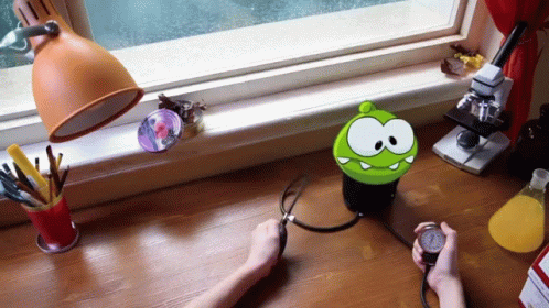 A Person Is Taking Their Blood Pressure With A Green Cartoon Character On The Desk GIF