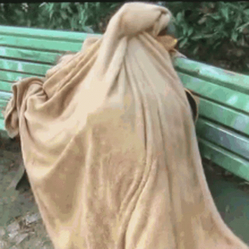 A Person Is Wrapped In A Blanket While Sitting On A Green Bench . GIF