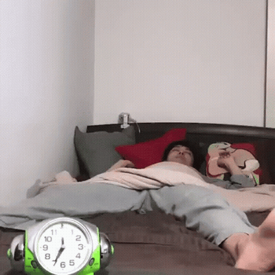 A Person Laying On A Bed Next To A Green Alarm Clock That Shows The Time As 7:15 GIF