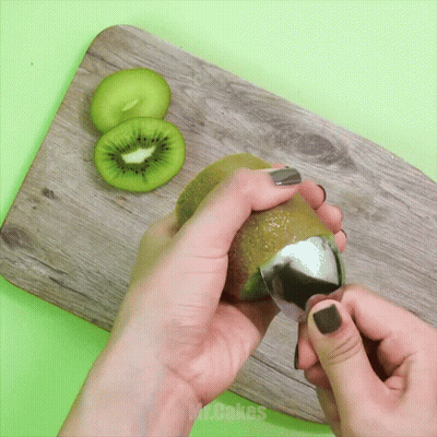 A Person Peeling A Kiwi With A Spoon On A Wooden Cutting Board GIF