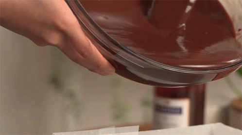 A Person Pouring Chocolate Into A Glass Bowl With A Bottle In The Background That Says ' Strawberry ' On It GIF