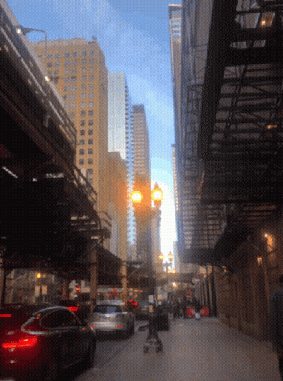 A Person Pushing A Stroller Down A City Street With Tall Buildings In The Background GIF