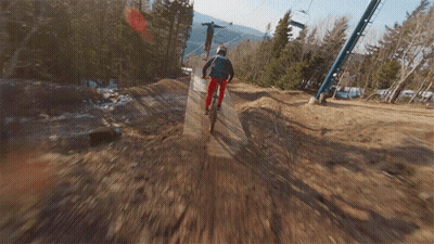 A Person Riding A Bike Down A Dirt Road With A Ski Lift In The Background GIF