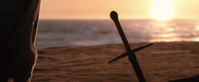 A Person Standing On A Beach With A Sword Laying On The Sand GIF