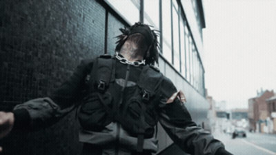 A Person Wearing A Black Jacket And A Mask With A Chain Around Their Neck Is Walking Down The Street GIF