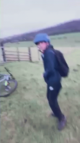 A Person Wearing A Blue Helmet And A Backpack Is Standing In A Field With A Bicycle In The Background . GIF