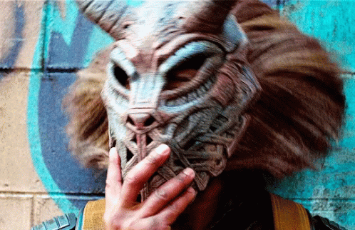 A Person Wearing A Mask With Horns Is Covering Their Mouth With Their Hand GIF
