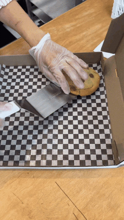 A Person Wearing Gloves Is Cutting A Chocolate Chip Cookie In A Box With A Spatula GIF