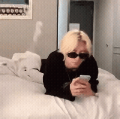 A Person Wearing Sunglasses Is Laying On A Bed Looking At Their Phone . GIF