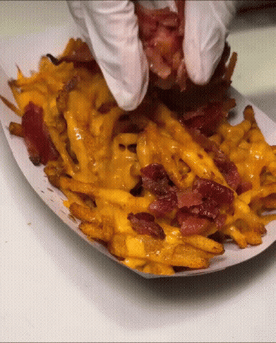 A Person Wearing White Gloves Holds A Piece Of Bacon Over French Fries GIF