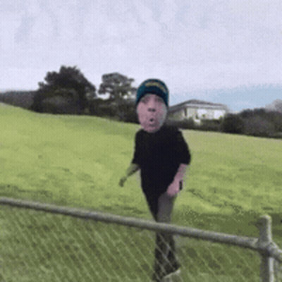 A Person With A Face On Their Head Is Walking Through A Grassy Field Behind A Chain Link Fence . GIF
