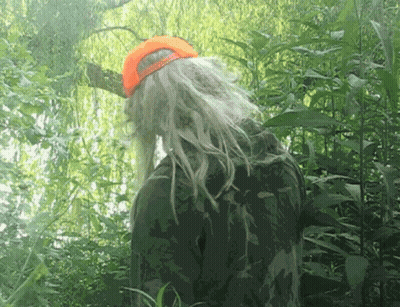 A Person With Long Blonde Hair And An Orange Hat Stands In The Woods GIF