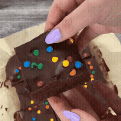 A Person With Purple Nails Is Holding A Piece Of Chocolate Cake With Sprinkles On Top . GIF