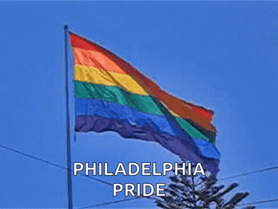 A Philadelphia Pride Flag Is Flying In The Wind Against A Blue Sky GIF