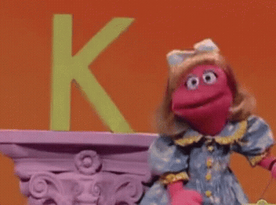 A Pink Puppet Is Standing In Front Of A Green Letter K On A Shelf . GIF