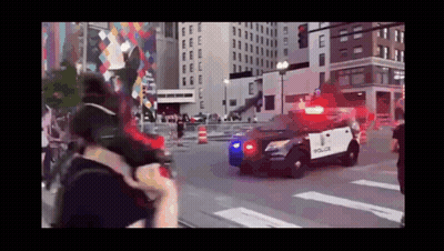 A Police Car Is Driving Down A City Street With A Crowd Behind It . GIF