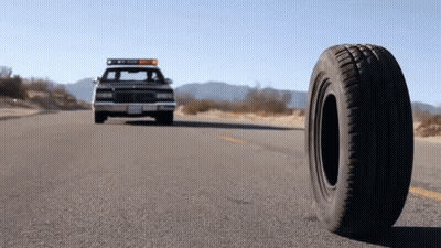 A Police Car Is Driving Down The Road With A Tire On The Side Of The Road GIF
