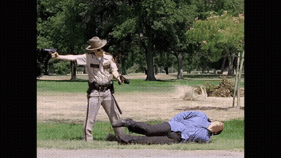 A Police Officer Is Pointing A Gun At A Man Laying On The Grass GIF