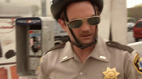 A Police Officer Wearing Sunglasses And A Helmet Stands In Front Of A Payphone GIF