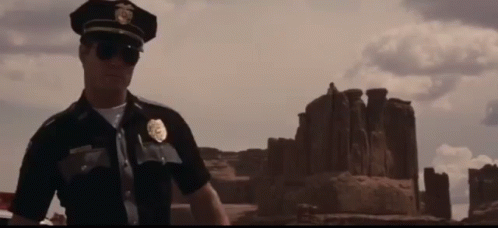 A Police Officer Wearing Sunglasses Is Standing In Front Of A Desert Landscape . GIF
