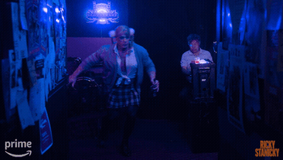 A Poster For Ricky Stanicky Shows A Man In A Plaid Skirt In A Dark Hallway GIF