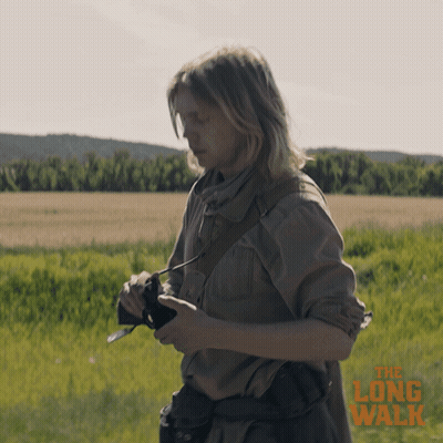 A Poster For The Long Walk Shows A Woman Holding A Camera In A Field GIF