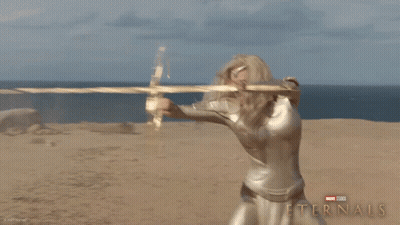 A Poster For The Movie Eternals Shows A Blonde Woman Holding A Golden Object GIF