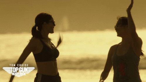 A Poster For Top Gun Maverick Shows Two Women Dancing On The Beach GIF