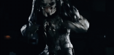 A Predator Is Standing In The Dark With Its Arms Outstretched And Looking At The Camera . GIF