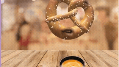 a pretzel is falling into a bowl of cheese sauce on a wooden table