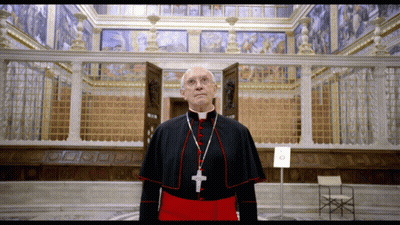 A Priest With A Cross Around His Neck Stands In Front Of A Building GIF
