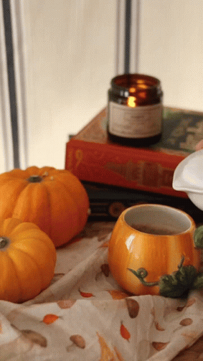 A Pumpkin Shaped Cup Sits On A Table Next To A Candle And Books GIF