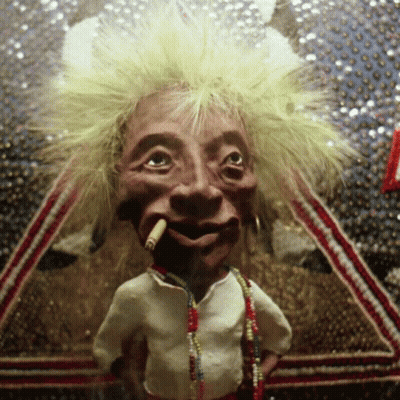 A Puppet With A Cigarette In His Mouth Is Wearing A White Shirt And Necklaces GIF