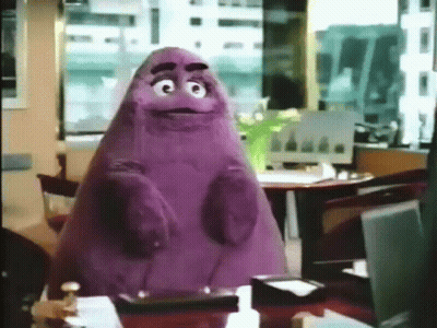A Purple Cartoon Character Is Sitting At A Table In An Office GIF