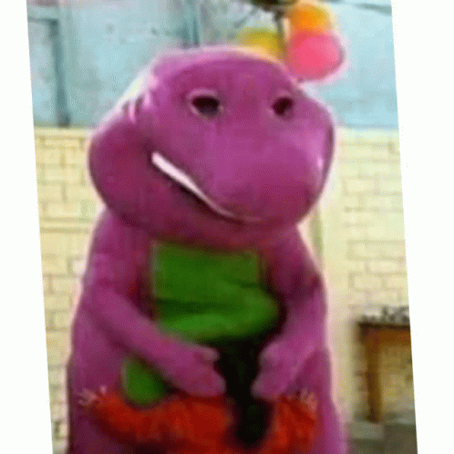 A Purple Dinosaur With A Green Tail Is Smiling In Front Of A Brick Wall GIF