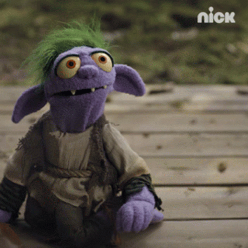 A Purple Puppet With Green Hair And Orange Eyes Is Sitting On A Wooden Surface With The Nick Logo In The Background GIF