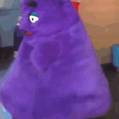 A Purple Stuffed Animal With A Blue Eye Is Sitting In A Room . GIF