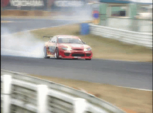 A Red Car Is Drifting On A Race Track With Smoke Coming Out Of The Tires GIF
