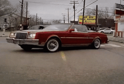 A Red Car Is Driving Down A Street In Front Of A Sign That Says We Buy Ugly Houses GIF