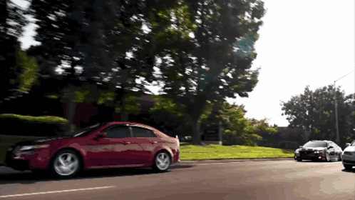 A Red Car Is Driving Down A Street Next To A Black Car GIF