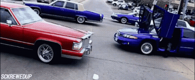 A Red Car Is Driving Down A Street Next To A Blue Car With Its Hood Open GIF
