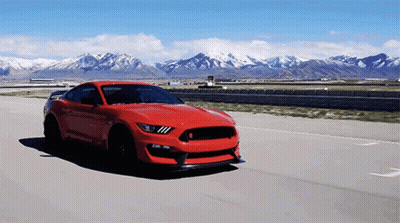 A Red Ford Mustang Is Driving Down A Road With Mountains In The Background GIF