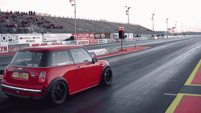 A Red Mini Cooper Is Driving Down A Race Track With A Lucas Oil Sign Behind It GIF