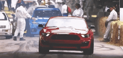 A Red Mustang Is Driving Down A Street With A Crowd Of People Behind It . GIF