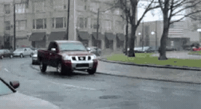 A Red Truck Is Driving Down A Wet Street In A City . GIF
